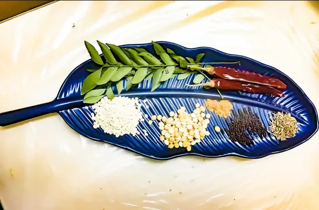 Auf einer blauen, blattförmigen Schale liegen ein Zweig Curryblätter, getrocknete rote Chilischoten sowie kleine Häufchen heller Linsen, Senfsaat und weiterer Samen.